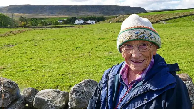 Margaret Bradshaw smiling at the camera. Behind it is a field, a farm and hills in the valley of Teesdale