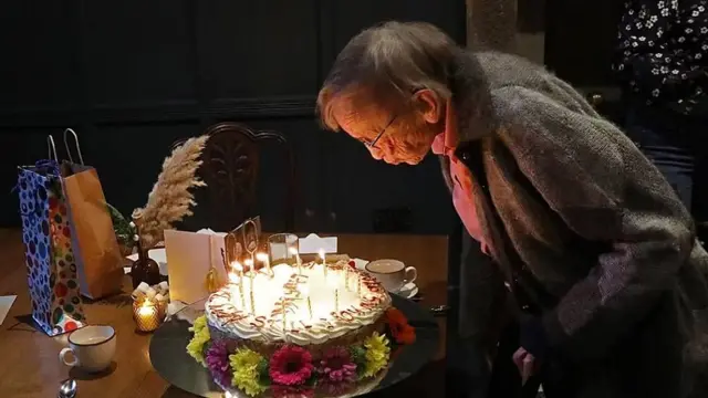 Margaret Bradshaw blows the candles in a large birthday cake covered in cream and decolarated with flowers